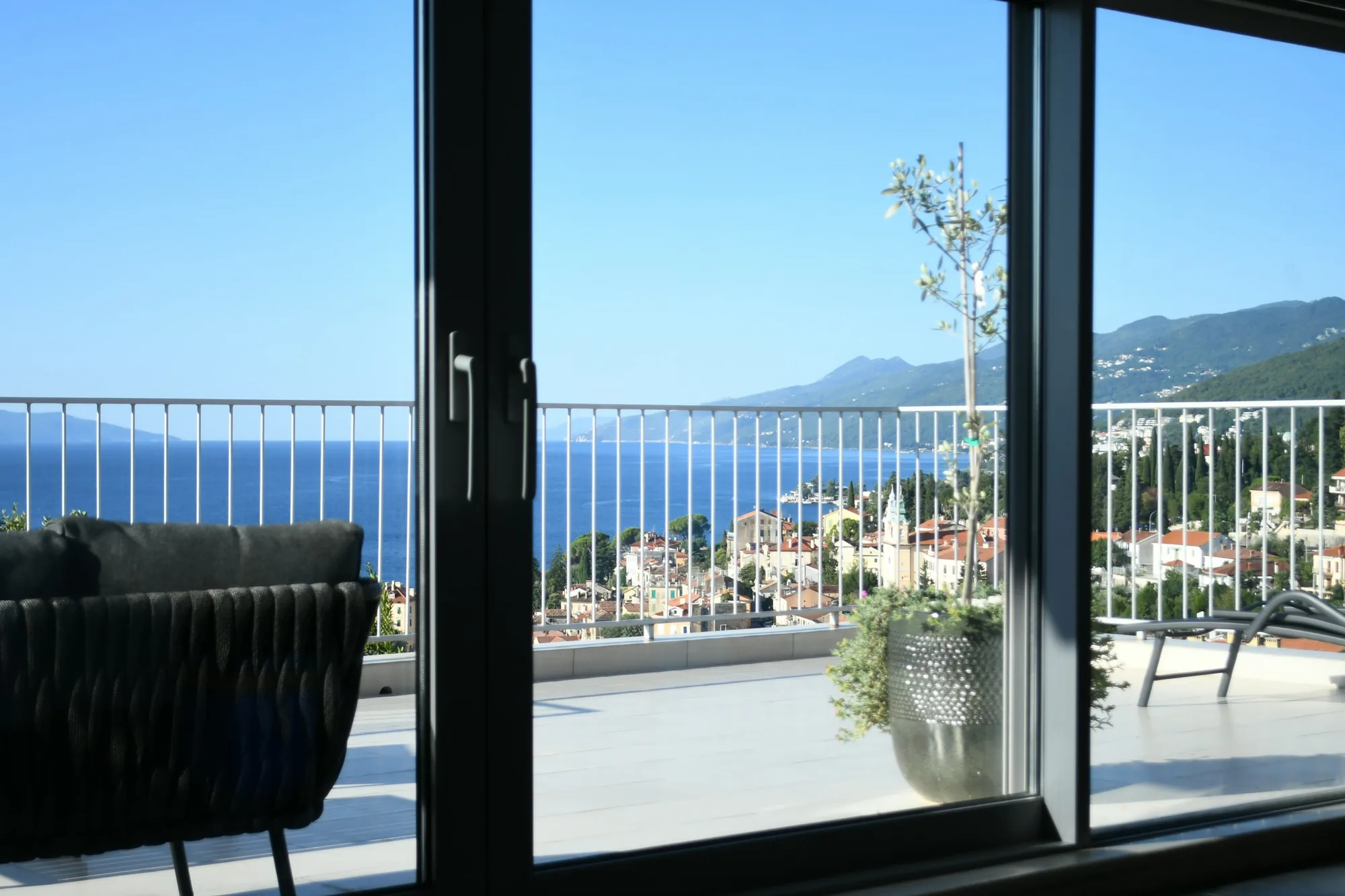 Baie vitrée d’un salon qui donne sur une terrasse, la mer et une petite ville de la côte